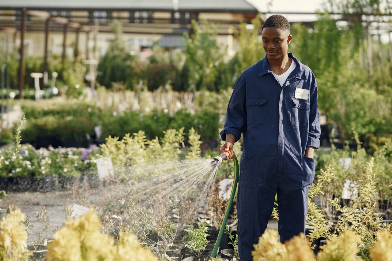 our-experience A smiling gardener in coveralls waters plants in a sunny outdoor nursery.