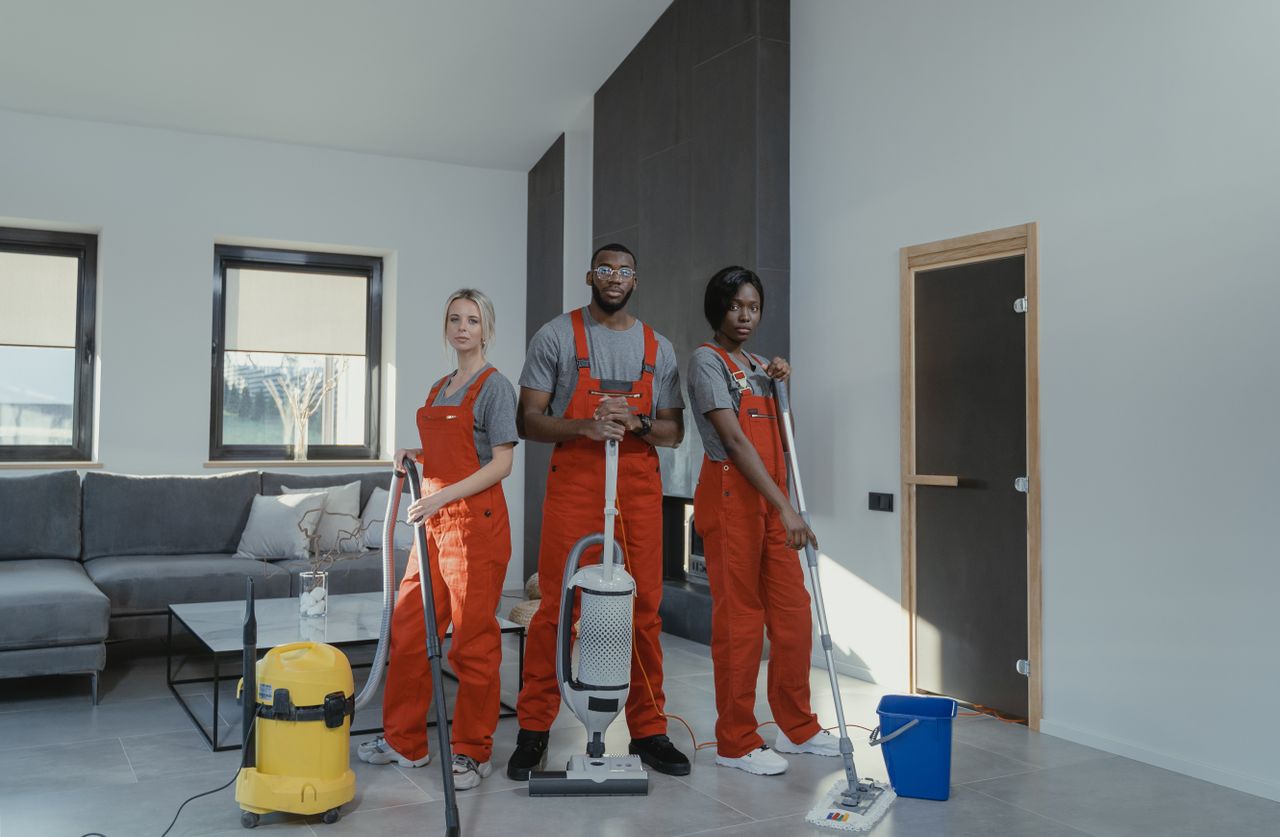 ours-journey Diverse cleaning team in red uniforms ready to sanitize a modern living room with vacuum and mop.