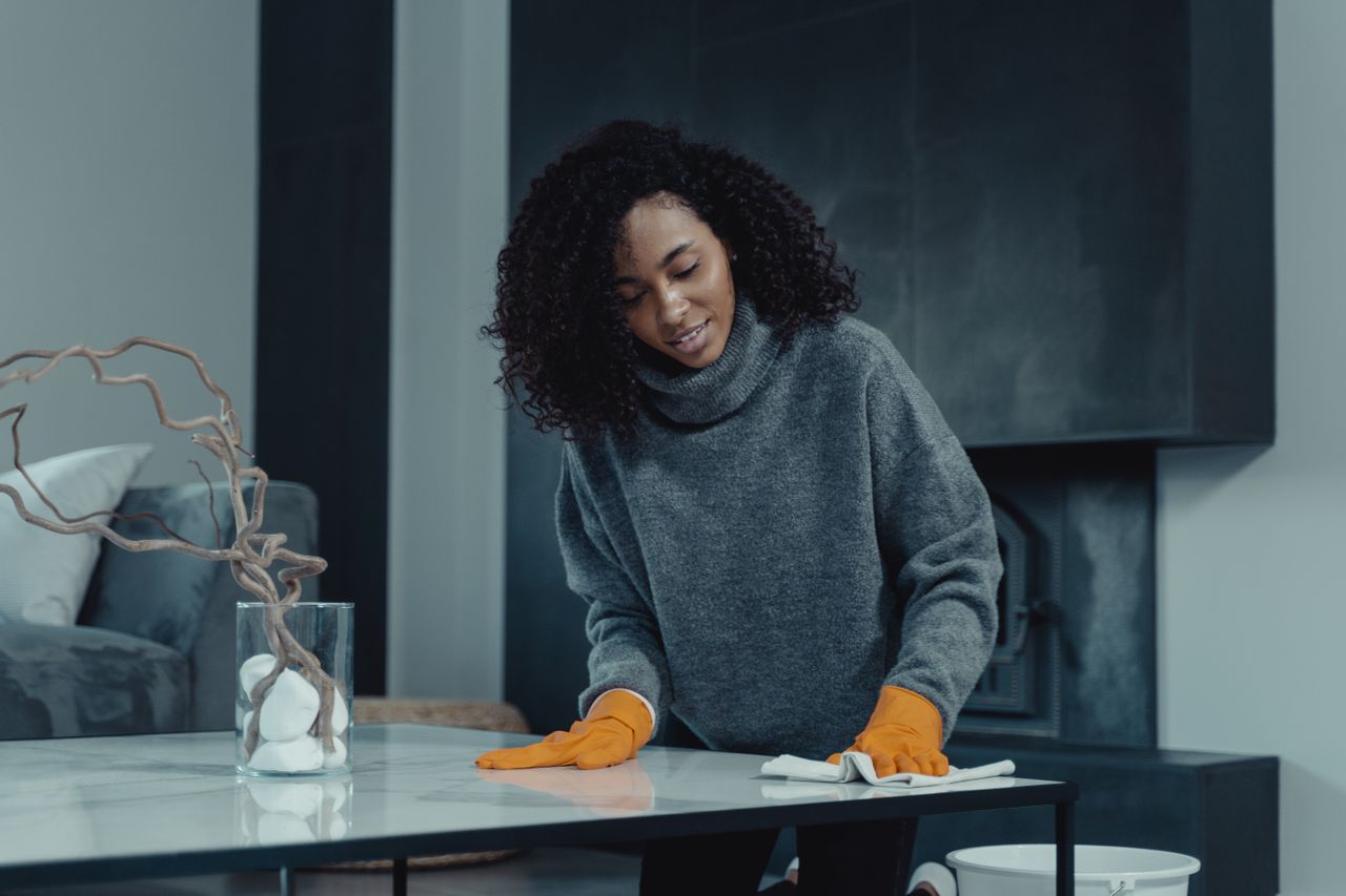 contact-img African American woman cleaning a table with orange gloves in a cozy, modern living room setting.